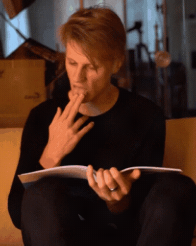 A Man Sitting On A Couch Reading A Book With His Hand On His Mouth GIF
