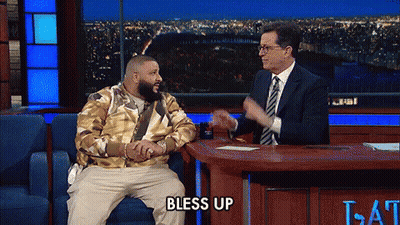 A Man Sitting On A Couch Talking To A Man In A Suit And Tie With The Words Bless Up On The Table GIF