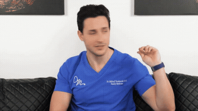 A Man Sitting On A Couch Wearing A Blue Scrub Top That Says Dr. Michał Tuchinski GIF