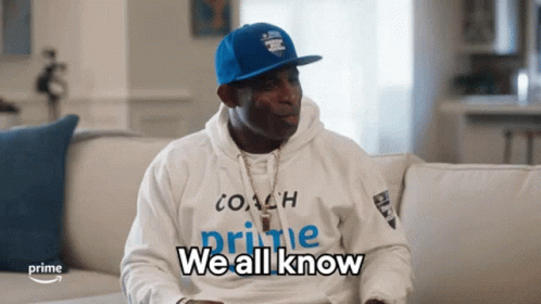 A Man Sitting On A Couch Wearing A White Hoodie That Says Coach Prime We All Know GIF