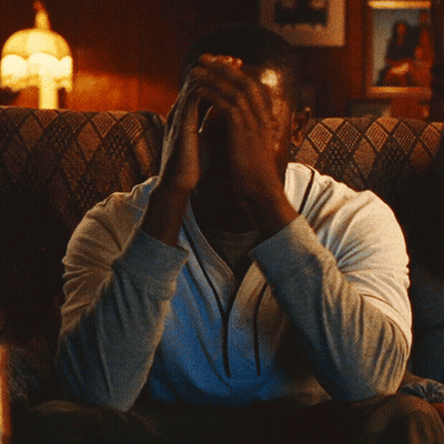 A Man Sitting On A Couch With His Hand On His Face GIF