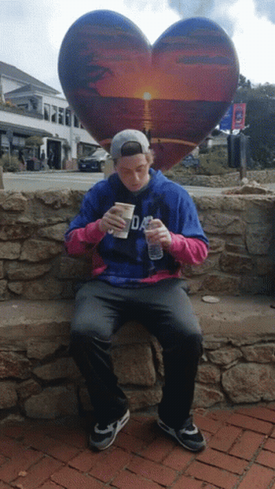 A Man Sitting On A Stone Wall With A Heart Shaped Sculpture In The Background GIF