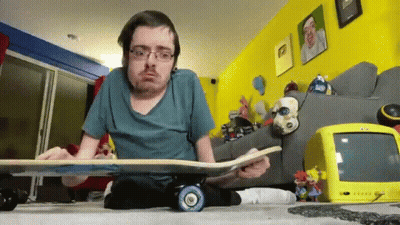 A Man Sitting On The Floor Holding A Skateboard With A Yellow Tv Behind Him GIF