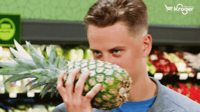 A Man Smells A Pineapple In A Grocery Store With A Kroger Logo In The Background GIF