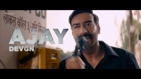 A Man Smoking A Cigarette In Front Of A Sign That Says A Jay Devgn GIF