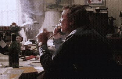 A Man Smoking A Cigarette While Talking On A Phone With A Bottle Of Wine In The Background GIF