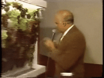 A Man Speaking Into A Microphone In Front Of A Display Of Plants GIF