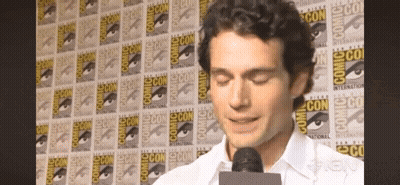 A Man Speaking Into A Microphone In Front Of A Wall That Says Comic Con On It GIF