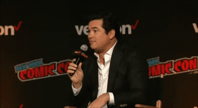 A Man Speaking Into A Microphone In Front Of A Wall That Says New York Comic Con GIF