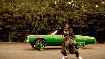 A Man Squatting Next To A Green Car With A Purple Stripe GIF