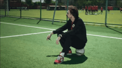 A Man Squatting On A Soccer Ball On A Field With A Team In The Background GIF