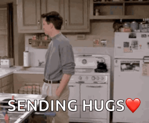 A Man Standing In A Kitchen With The Words " Sending Hugs " Above Him GIF