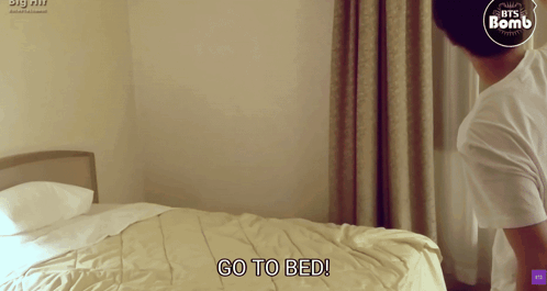 A Man Standing In Front Of A Bed With The Words Go To Bed GIF