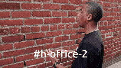 A Man Standing In Front Of A Red Brick Wall With The Hashtag # H-Office-2 GIF