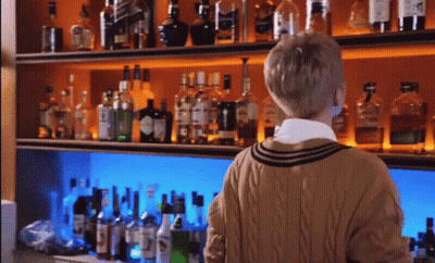 A Man Standing In Front Of A Shelf Full Of Liquor Bottles Including A Bottle Of Gin GIF