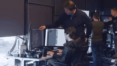 A Man Standing Next To A Man Sitting At A Desk Looking At A Computer Screen GIF