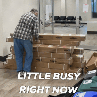 A Man Standing Next To A Pile Of Cardboard Boxes With The Words Little Busy Right Now Above Him GIF