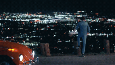 A Man Standing Next To An Orange Car Looking At A City GIF