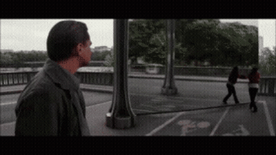 A Man Standing On A Bridge Looking At A Woman Walking On The Sidewalk GIF