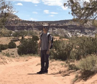 A Man Standing On A Dirt Road Wearing Sunglasses And A Hat GIF