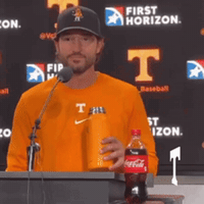A Man Stands At A Podium Holding A Coca Cola Bottle And A Water Bottle GIF