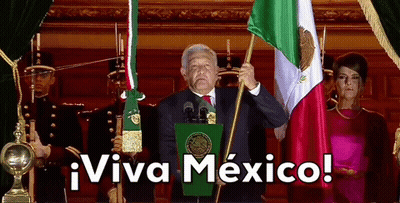 A Man Stands At A Podium Holding A Mexican Flag With The Words Viva Mexico Above Him GIF