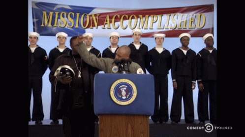 A Man Stands At A Podium In Front Of A Mission Accomplished Banner GIF