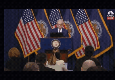 A Man Stands At A Podium In Front Of American Flags With Cnbc Live On The Screen Behind Him GIF