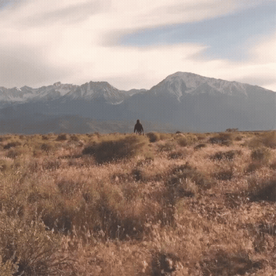 A Man Stands In A Field With Mountains In The Background GIF