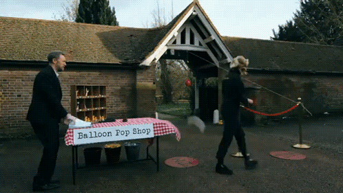 A Man Stands In Front Of A Balloon Pop Shop Table GIF