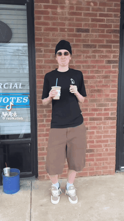 A Man Stands In Front Of A Brick Building Holding A Cup Of Coffee GIF