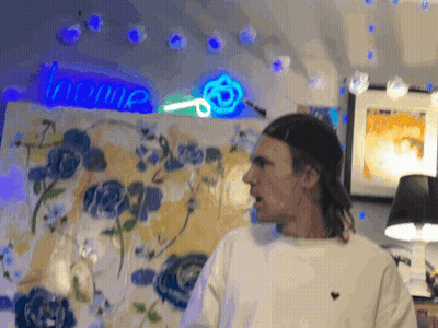 A Man Stands In Front Of A Painting With A Neon Sign That Says Home Behind Him GIF