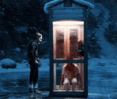 A Man Stands In Front Of A Phone Booth With A Woman Sitting Inside GIF