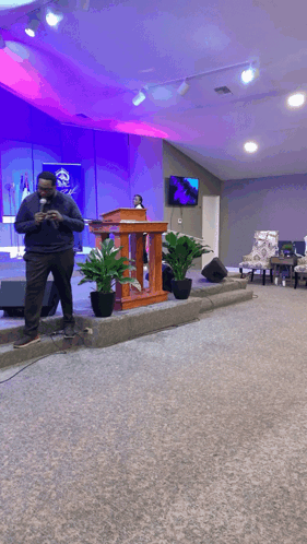 A Man Stands In Front Of A Podium In A Church With Potted Plants GIF