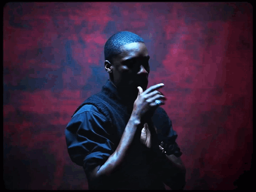 A Man Stands In Front Of A Red Wall With His Hands Folded GIF