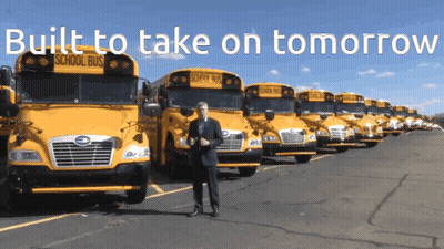 A Man Stands In Front Of A Row Of School Buses With The Words Built To Take On Tomorrow Above Him GIF