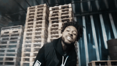 A Man Stands In Front Of A Stack Of Wooden Pallets With The Letter T On His Shirt GIF