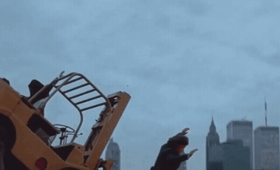 A Man Stands In Front Of A Yellow Forklift In A City GIF