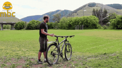 A Man Stands Next To A Bicycle In A Grassy Field With Mountains In The Background And The Word Mbc In The Corner GIF