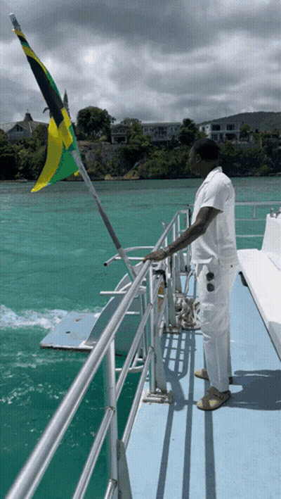 A Man Stands On A Boat With A Flag Flying In The Background GIF