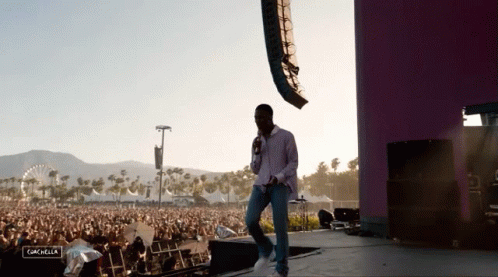 A Man Stands On A Stage With A Microphone In Front Of A Crowd At A Music Festival And The Word Coachella Is Visible GIF