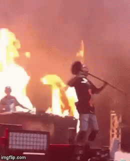 A Man Stands On A Stage With A Microphone In Front Of A Fire GIF