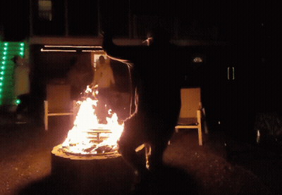 A Man Stands On A Stump In Front Of A Fire At Night GIF