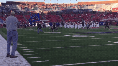 A Man Stands On The Sideline Of A Football Field Watching A Marching Band GIF