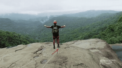 A Man Stands On Top Of A Rocky Cliff With His Arms Outstretched In Front Of A Lush Green Forest .. GIF