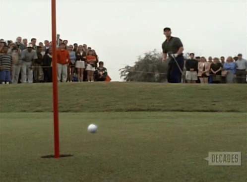 A Man Swings A Golf Club At A Golf Ball On A Golf Course With People Watching GIF