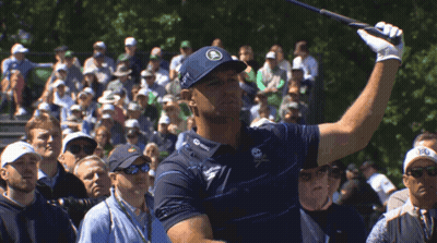 A Man Swings A Golf Club In Front Of A Crowd That Includes A Man Wearing A Hat That Says R On It GIF