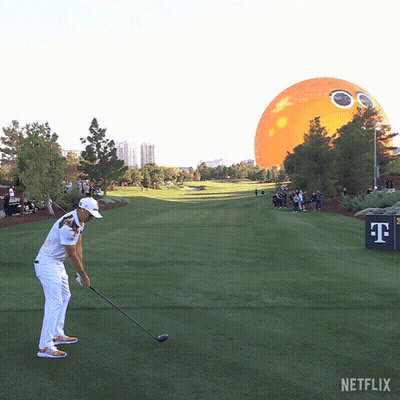A Man Swings A Golf Club On A Golf Course With An Orange Balloon In The Background GIF