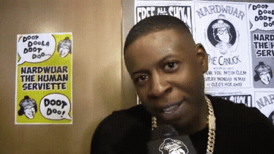 A Man Talking Into A Microphone In Front Of Posters That Say Nardwuar GIF