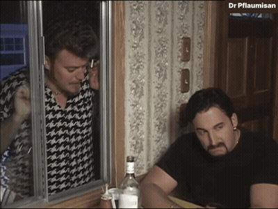 A Man Talking On A Cell Phone Next To A Man Sitting At A Table With A Bottle Of Vodka GIF